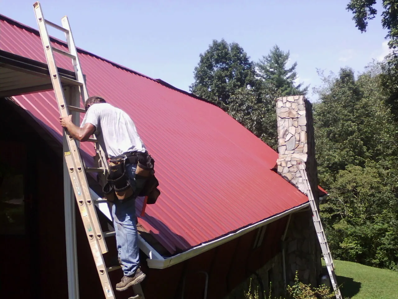 Roofer on a ladder beside a red metal roof and stone chimney — Soffit & Fascia Repair by Roofing Brothers in Rocky Face
